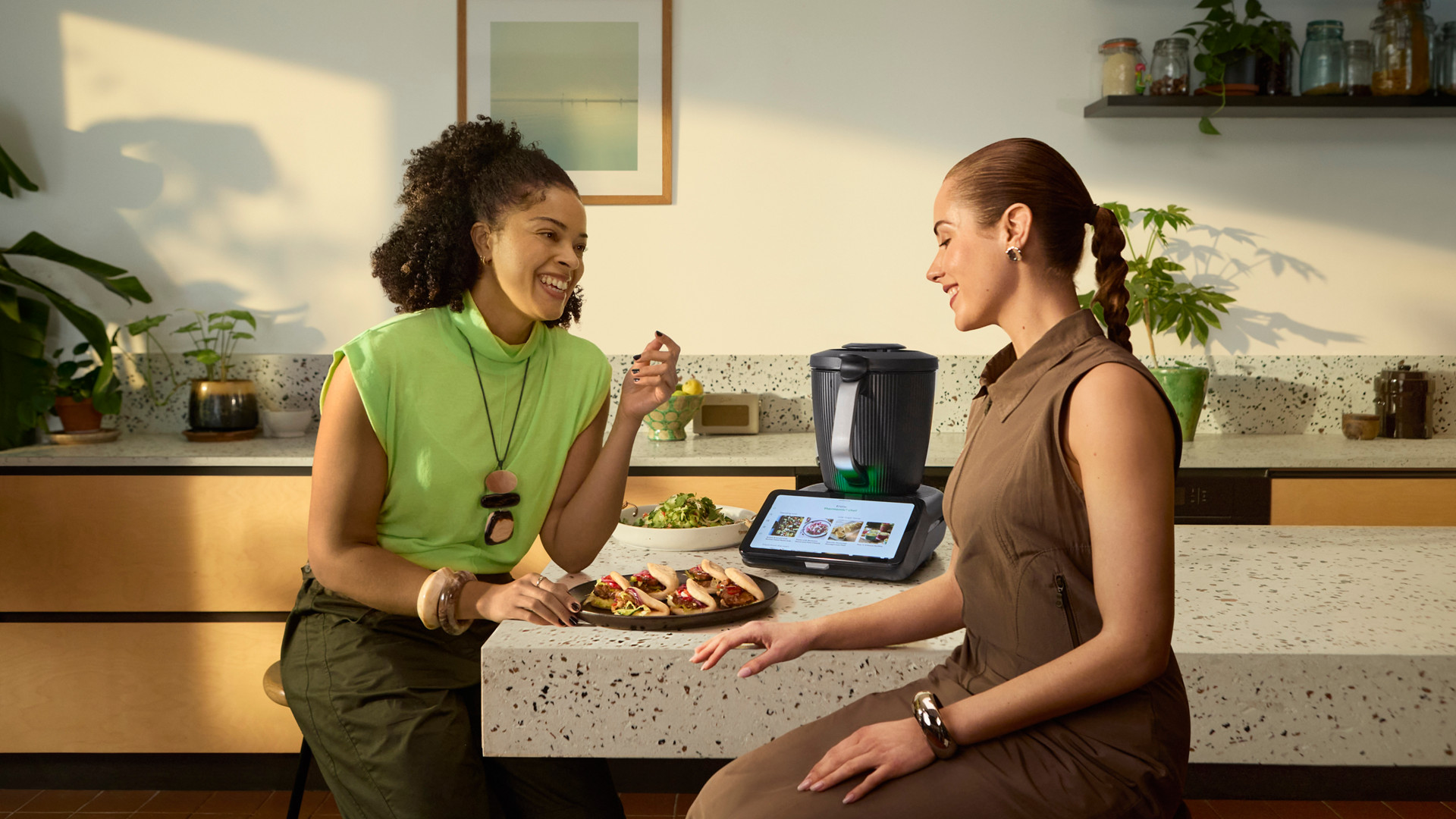 Two women sit at a kitchen counter with food and a Thermomix® TM7 on the countertop.