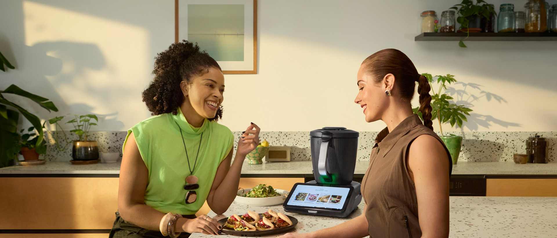 Two women sit at a kitchen counter with food and a Thermomix® TM7 on the countertop.