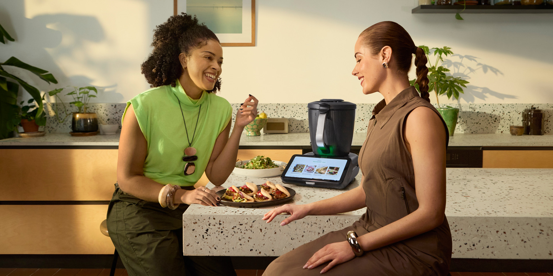 Two women sit at a kitchen counter with food and a Thermomix® TM7 on the countertop.