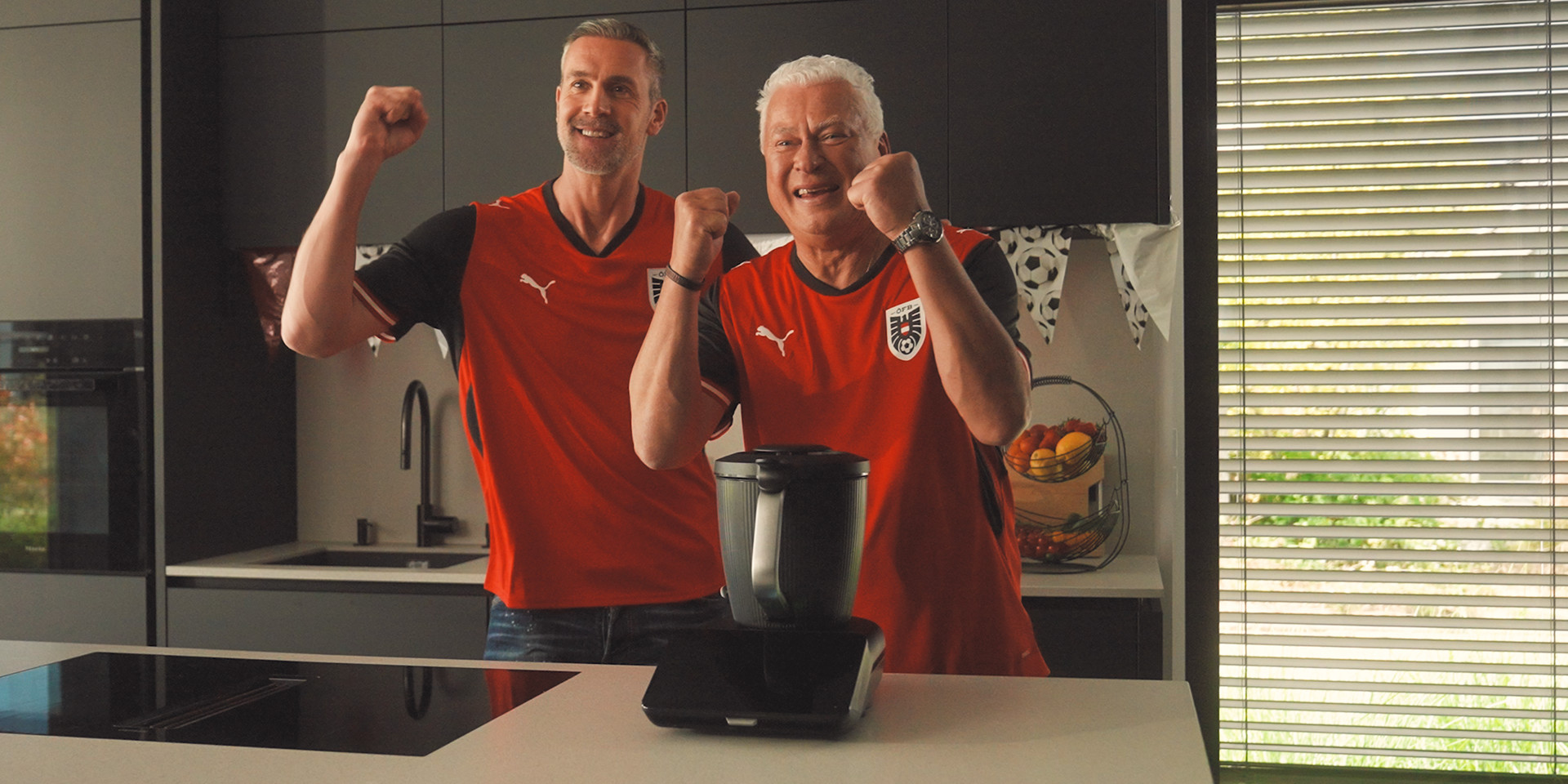 Zwei Personen in roten Österreich-Fußballtrikots jubeln in einer Küche mit einem Thermomix® TM7 auf der Arbeitsplatte.