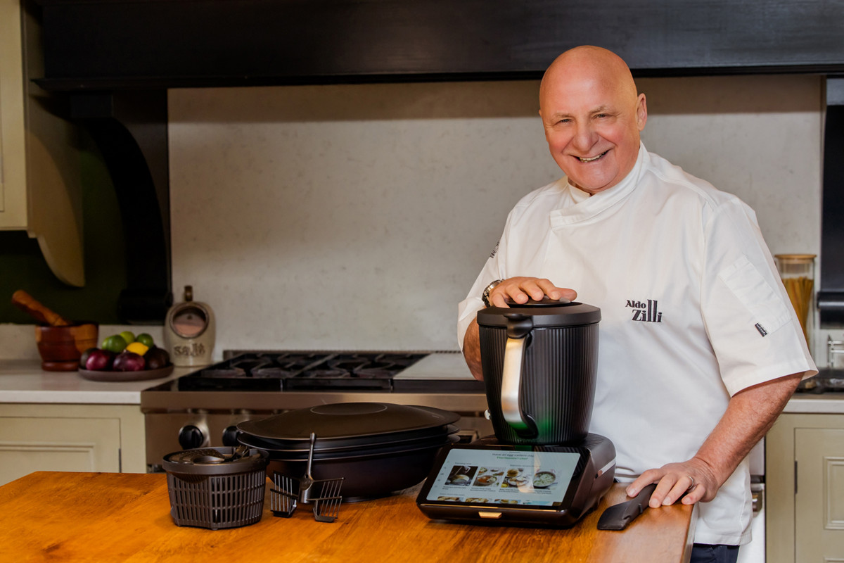 Aldo Zilli with Thermomix, smiling at the camera, his hand on the Thermomix, table in front of him with accessoires for the TM7