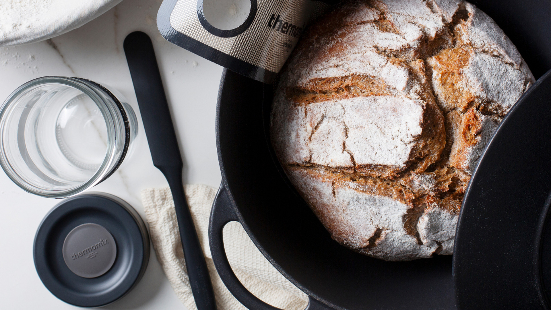 Frisch gebackenes Brot im schwarzen Topf, umgeben von Thermomix® Backzubehör auf bemehlter Arbeitsfläche.