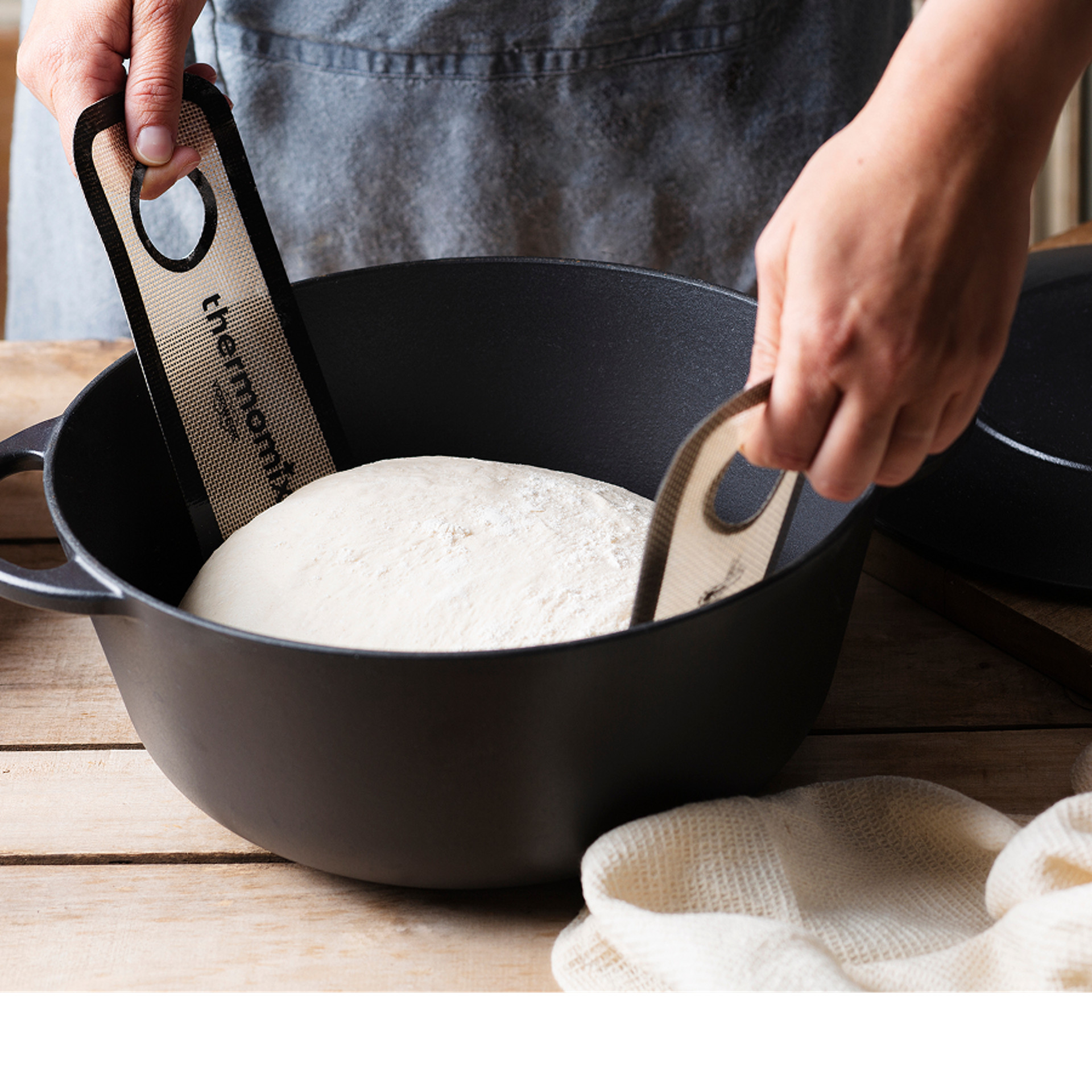 Hände heben Brotteig in einem schwarzen Gusseisentopf mit Thermomix® Teighebegurten auf einer Holzoberfläche.