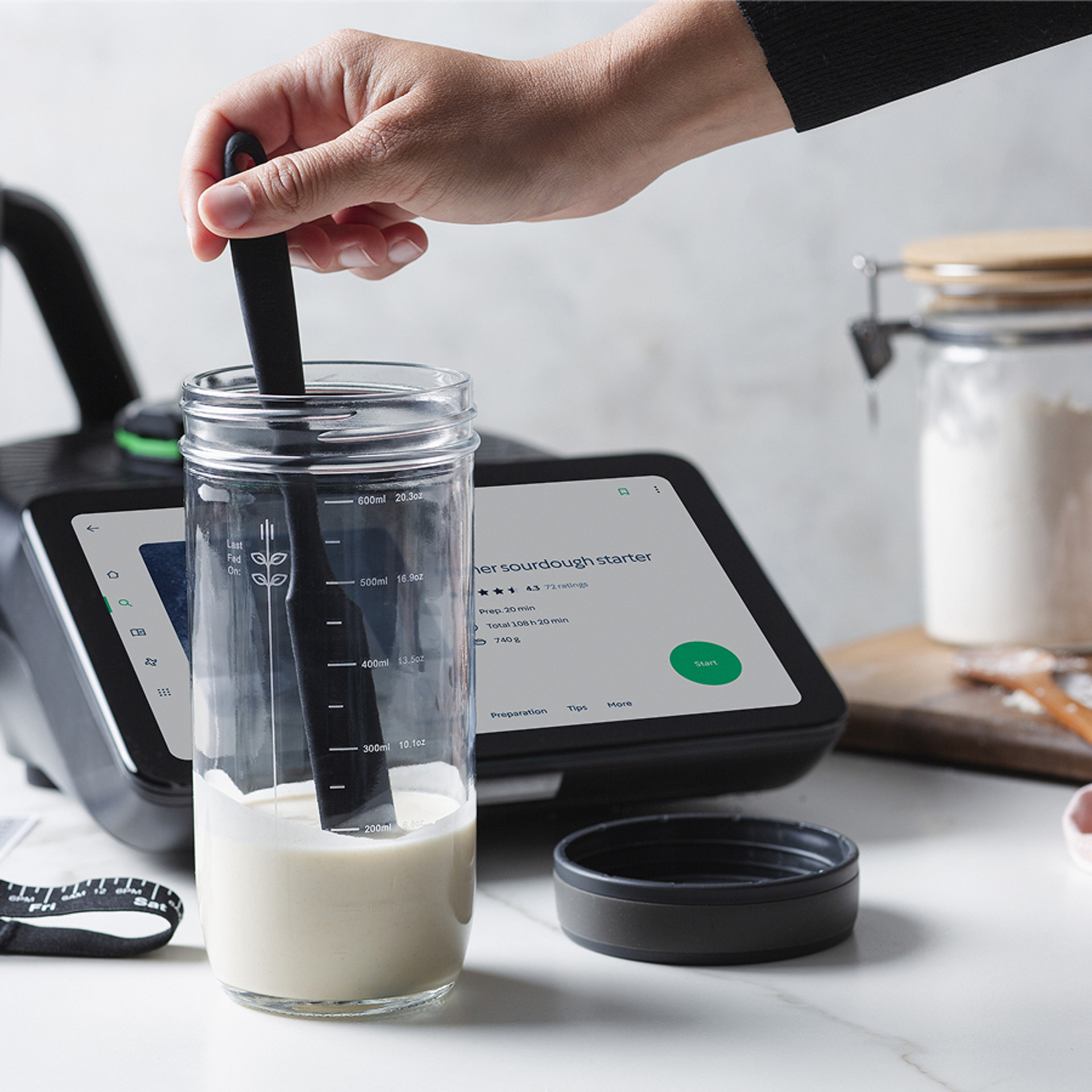 Hand mischt Sauerteigstarter in einem Glas neben Thermomix® TM7 mit Rezeptanzeige auf Touchscreen.