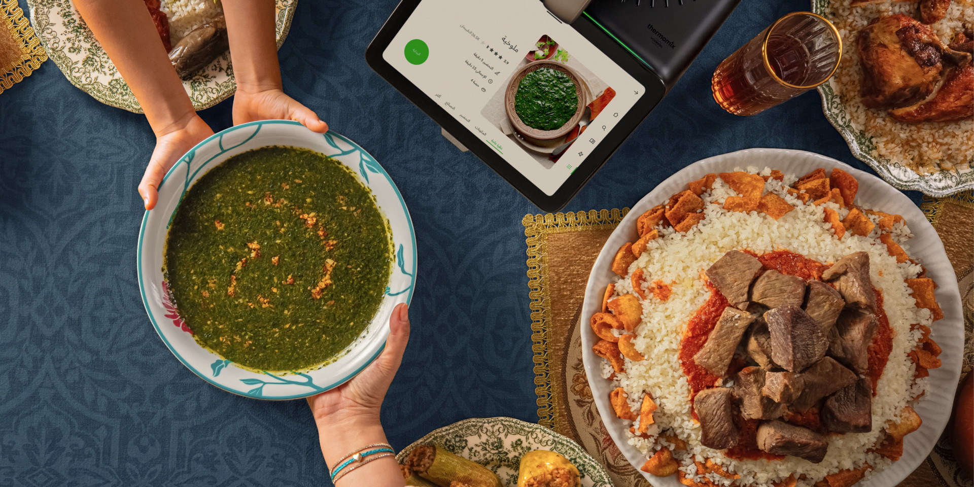 Overhead view of various Middle Eastern dishes on a blue tablecloth, with a Thermomix® TM7 showing a green soup recipe.