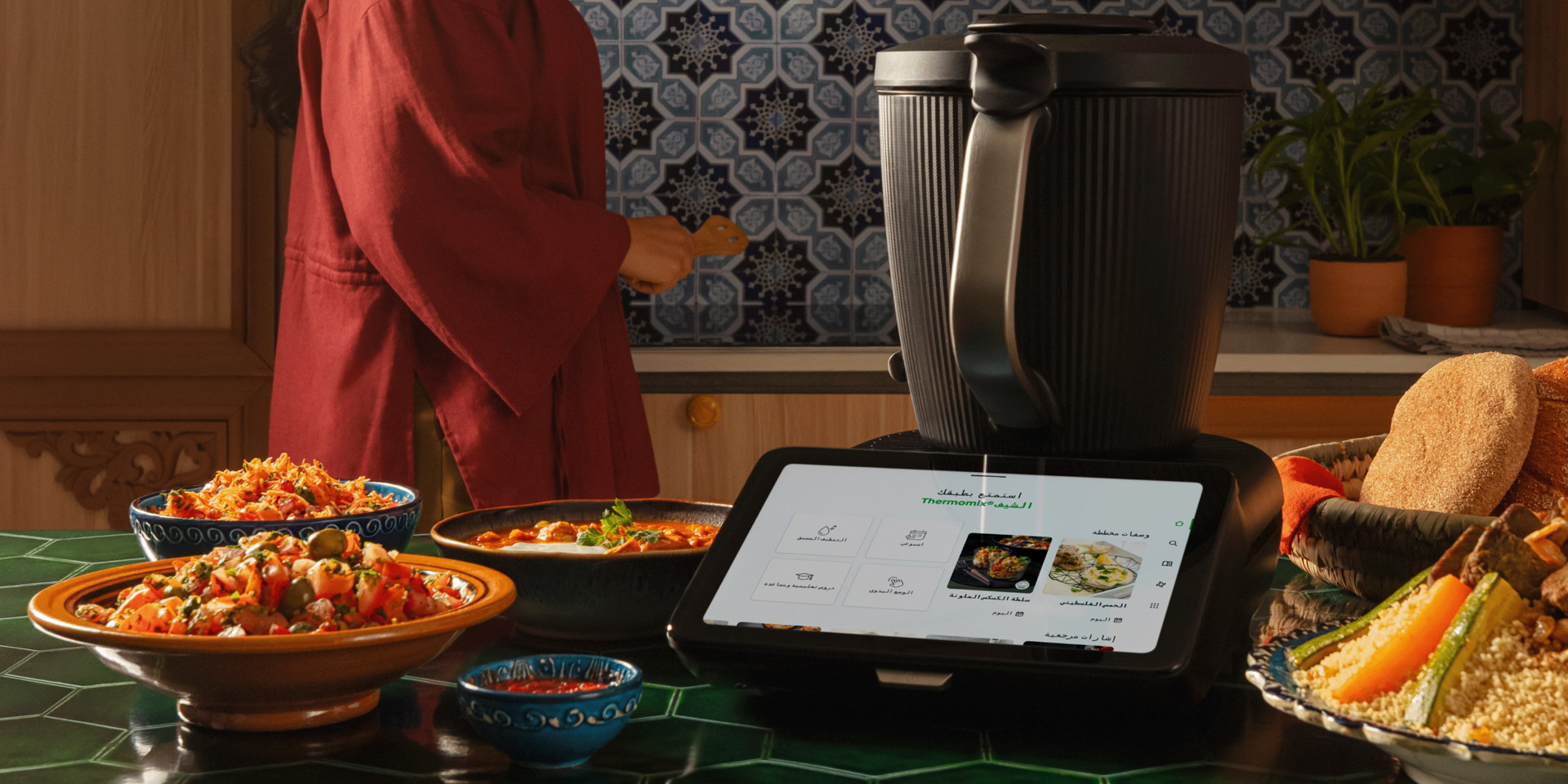 Thermomix® TM7 in a kitchen setting with prepared dishes on the table and a person in the background.