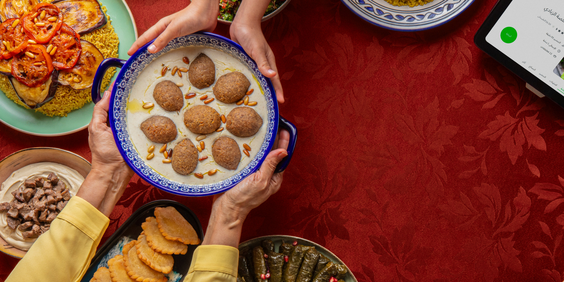 Shared meal with various Middle Eastern dishes and a Thermomix® TM6 recipe display on a red tablecloth.