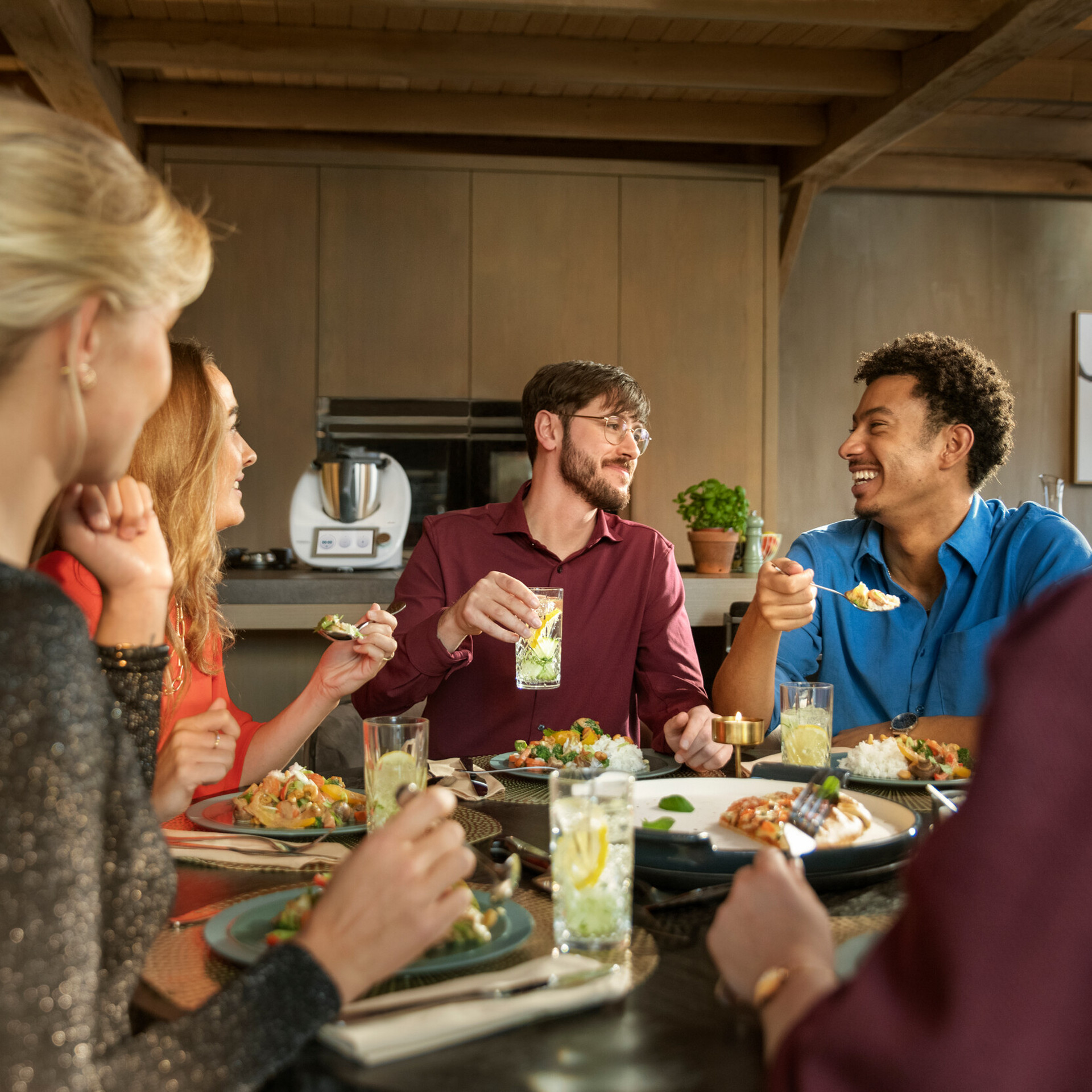 Grupo de personas cenando en una mesa con un Thermomix® TM6 visible en la cocina al fondo.