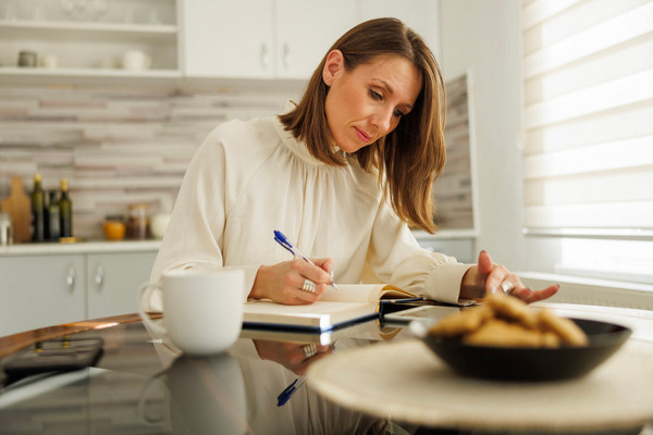 Mujer caucásica de mediana edad, empresaria, tomando notas en su agenda y usando una tableta mientras trabaja desde casa.