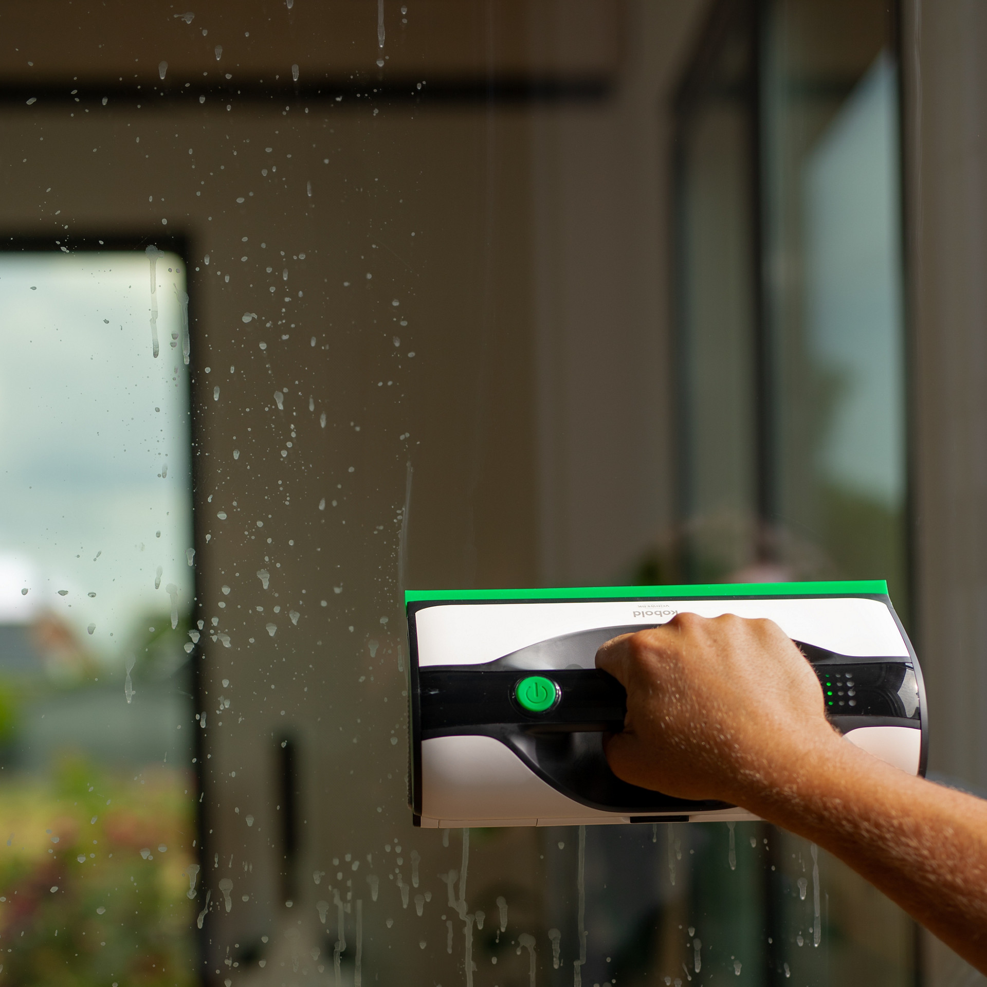 Main nettoyant une vitre avec le nettoyeur de vitres Kobold VG100+, visible à travers des traces savonneuses.