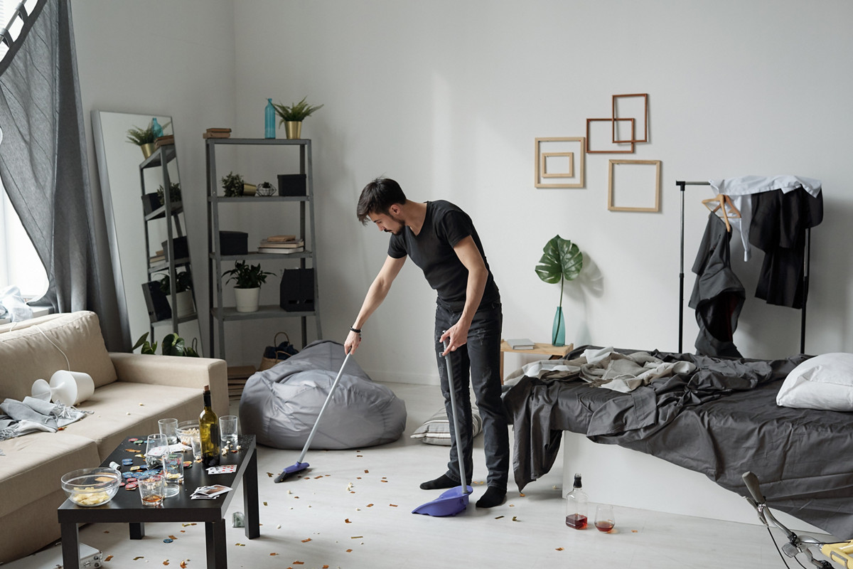 Young man sweeping floor at home after party: bottles, crumps and poker chips on floor