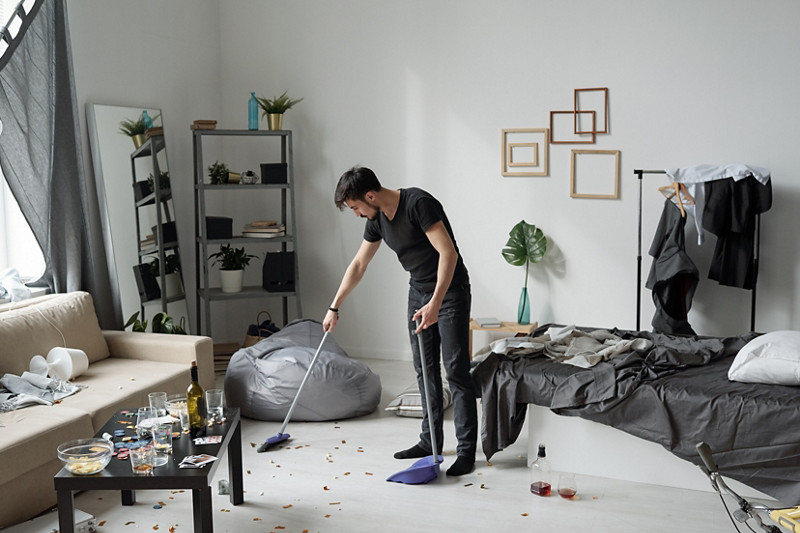 Jeune homme balayant le sol de sa maison après une fête : bouteilles, confettis et papiers