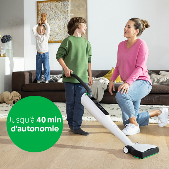 Enfant passe l aspirateur Kobold VK7 sur un sol en bois femme accroupie et autre enfant jouant au fond