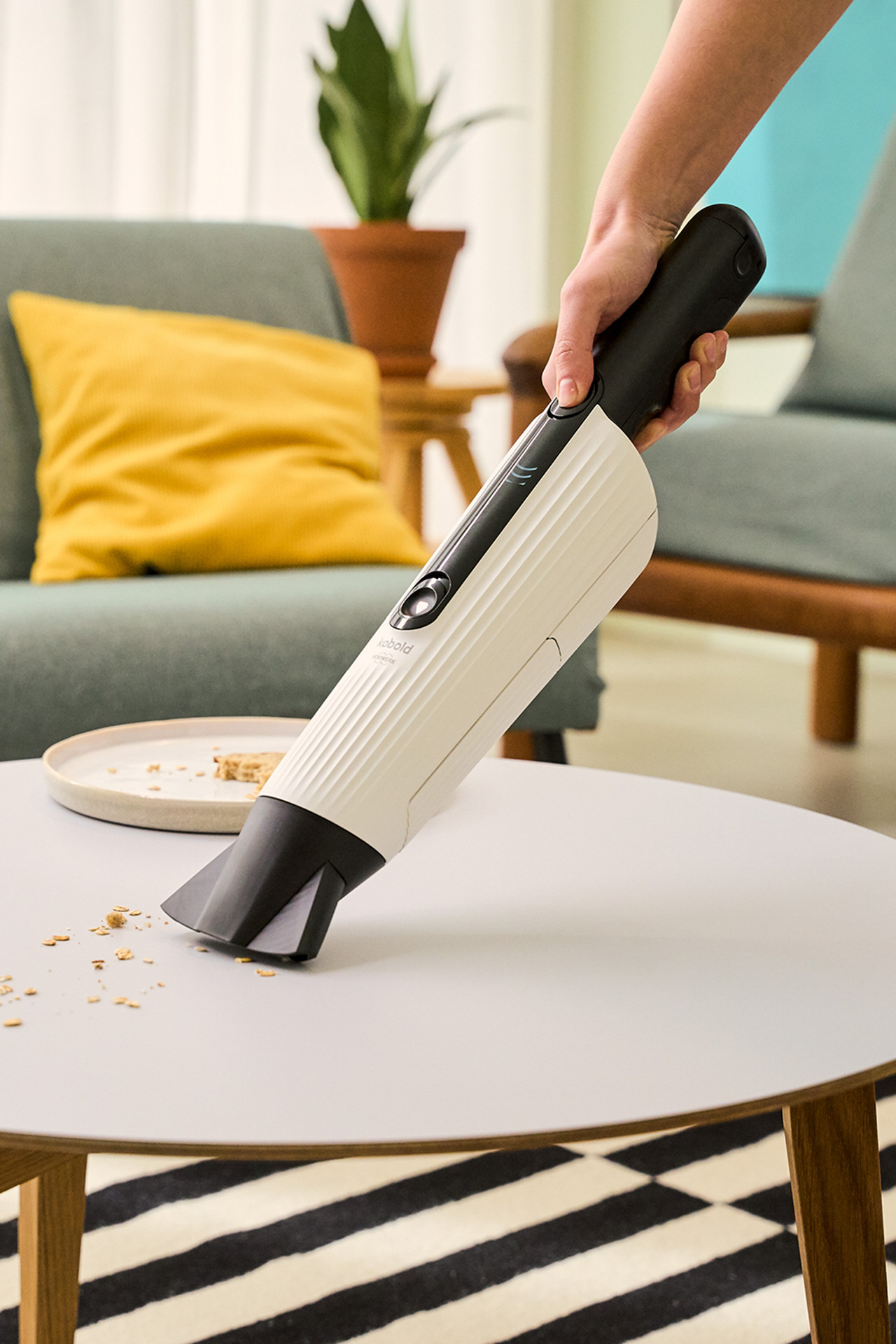 Une personne utilise l’aspirateur de table Kobold VM7 pour aspirer des miettes sur une table ronde dans un salon.
