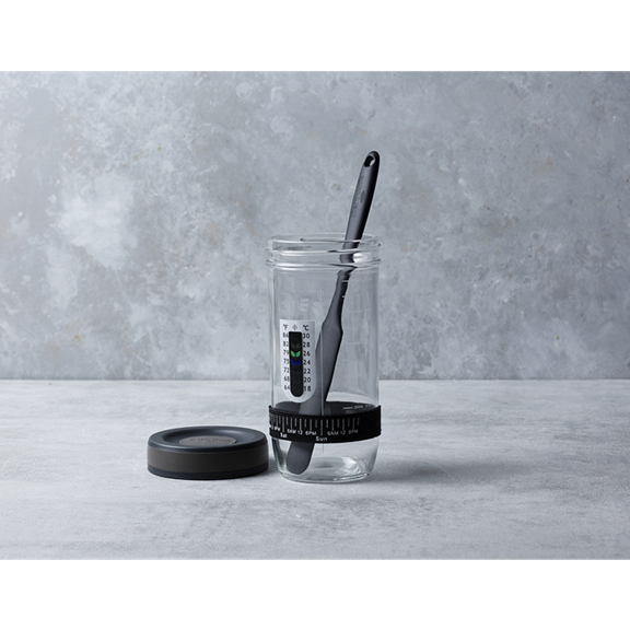 Bocal en verre avec couvercle noir spatule et repères de mesure sur surface grise et fond en pierre