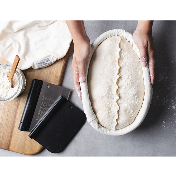Mains façonnant une pâte dans un panier avec de la farine une corne et un linge Thermomix à côté