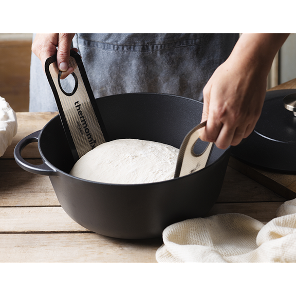 Mains soulevant une pâte à pain dans une cocotte noire avec des sangles Thermomix sur une table en bois
