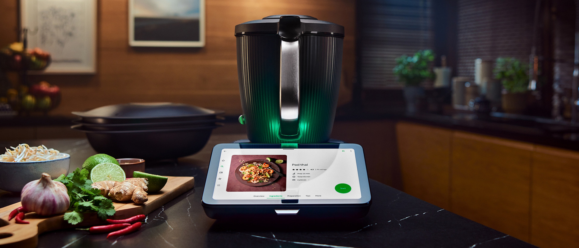 Thermomix® TM7 sur le plan de travail de cuisine, affichant une recette, entouré d’ingrédients frais et d’ustensiles.