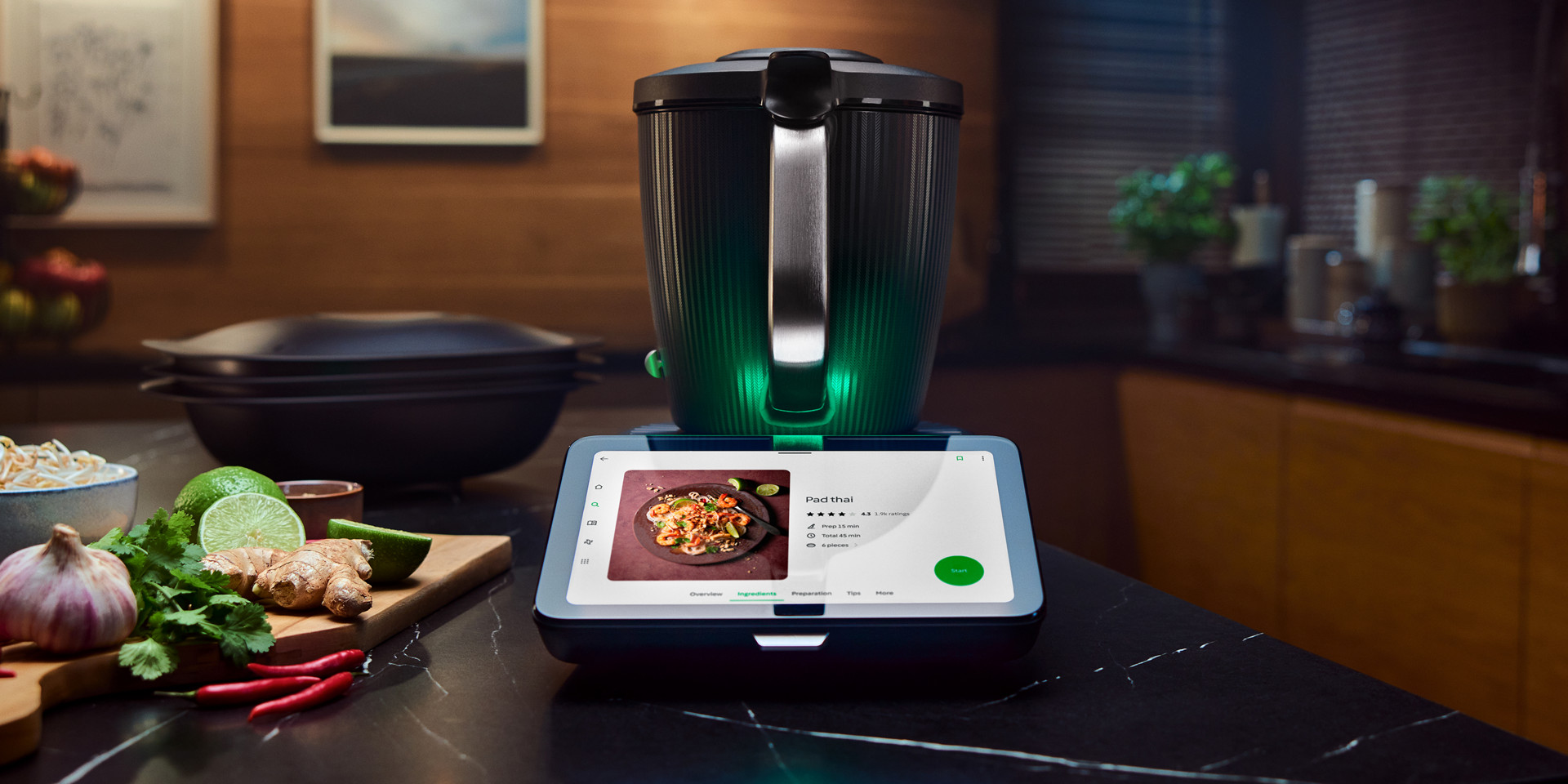 Thermomix® TM7 sur le plan de travail de cuisine, affichant une recette, entouré d’ingrédients frais et d’ustensiles.