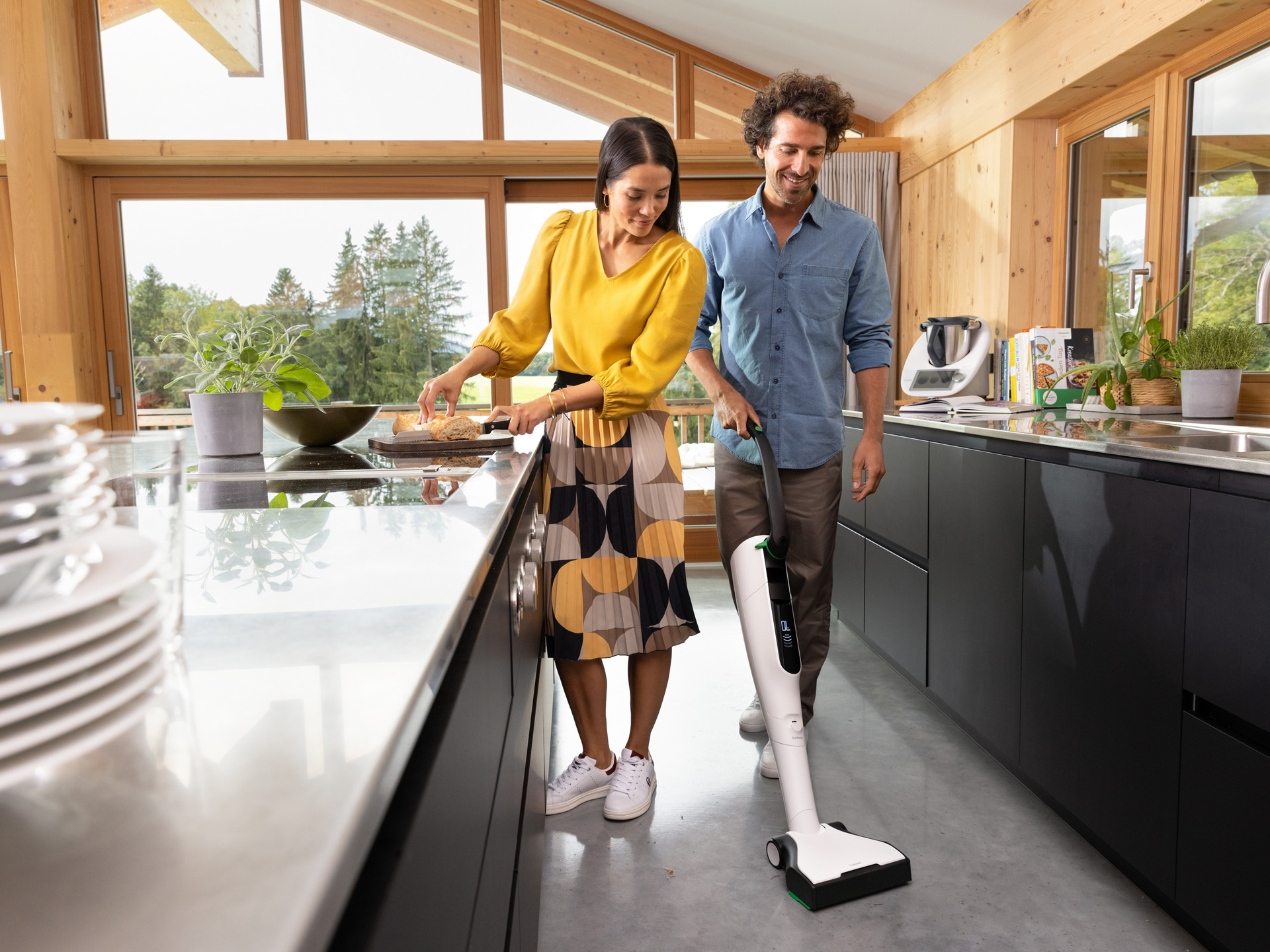 Homme passant l’aspirateur Kobold VK7 dans une cuisine moderne, Thermomix® TM6 sur le plan, femme cuisine.