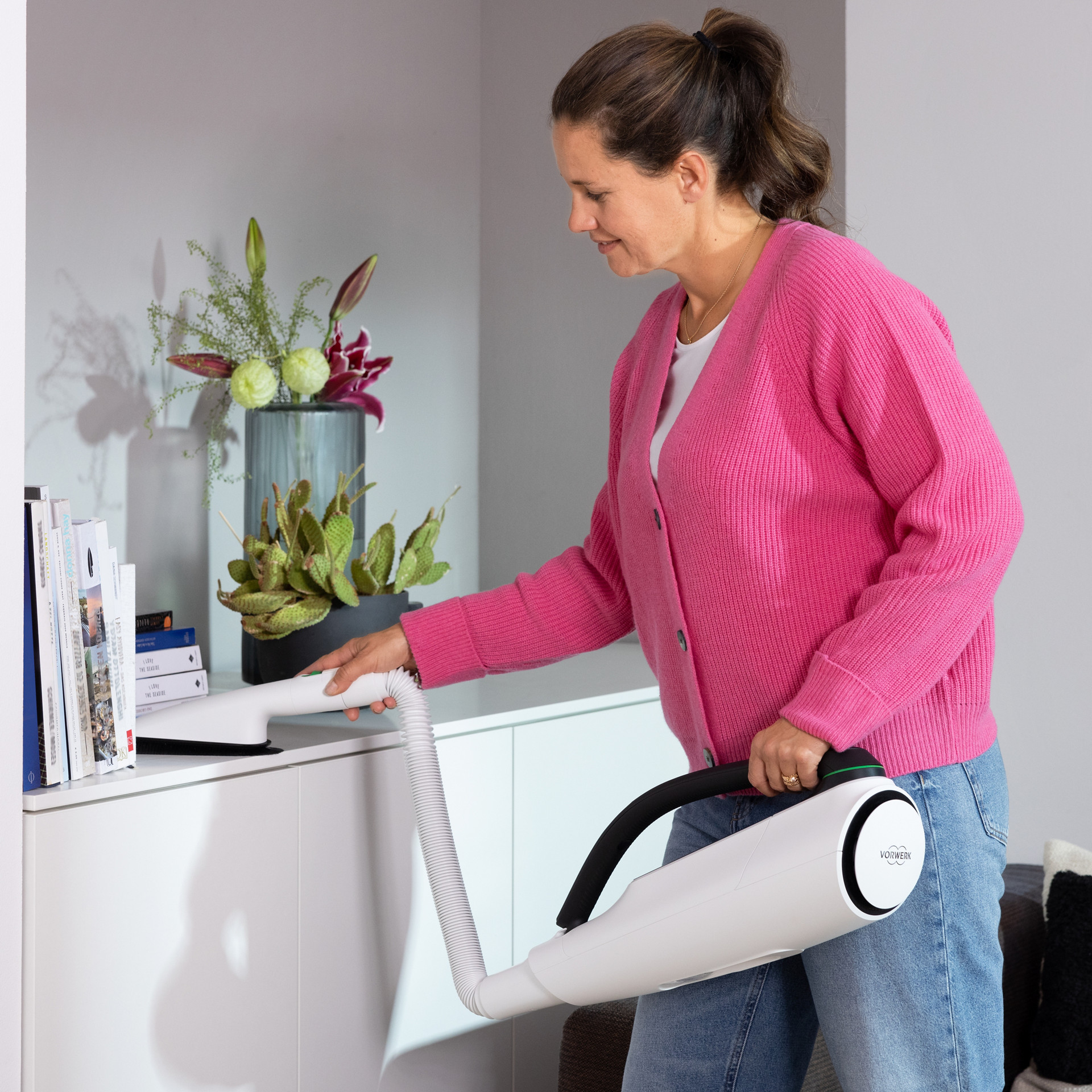 Personne passant l’aspirateur sur un meuble avec le Kobold VT300, près de livres et d’un vase de fleurs.