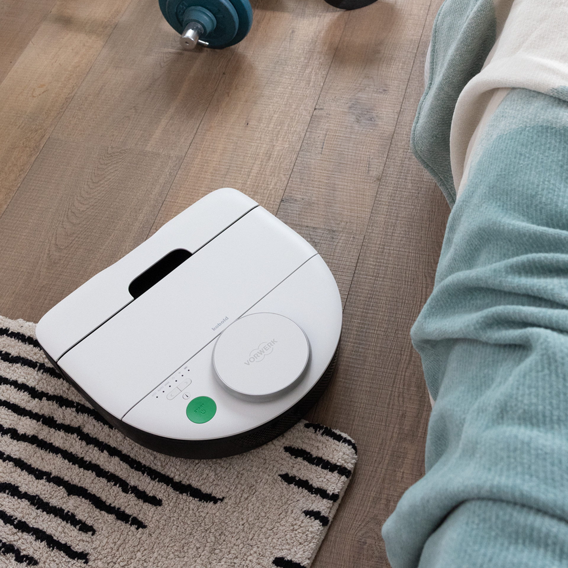 Robot aspirateur VR7 nettoie un sol en bois près d’un tapis, d’un lit et d’haltères.