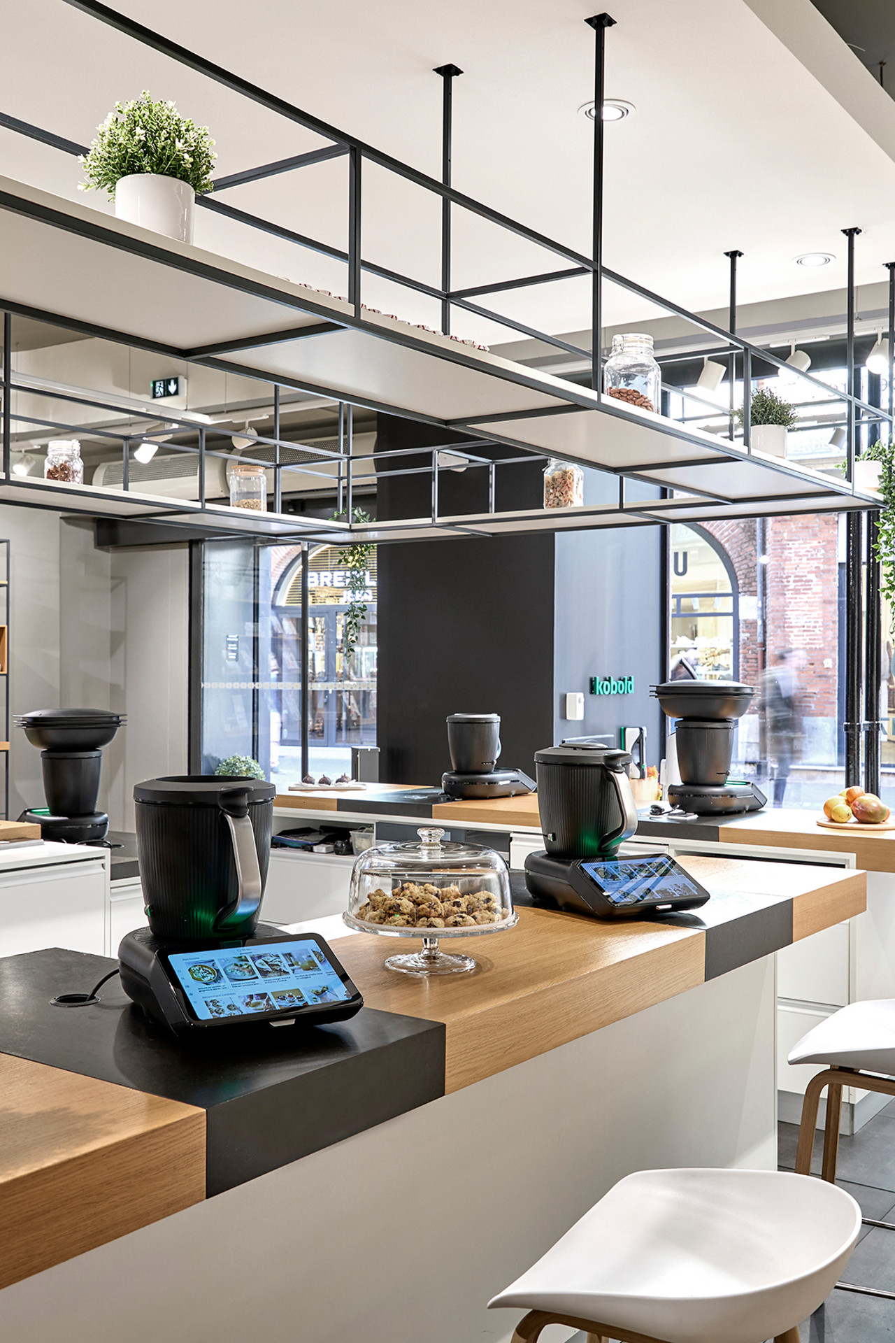 Showroom de cuisine moderne avec plusieurs Thermomix® TM7 sur les tables, tabourets et étagères d’ouvrages culinaires.