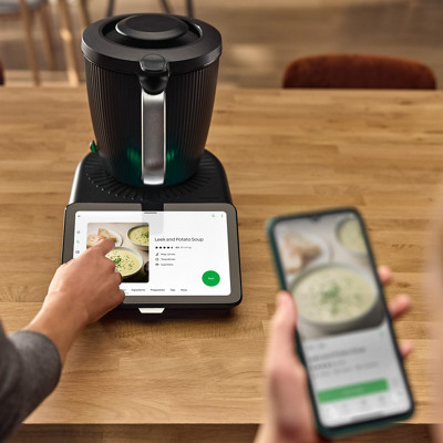 Le robot thermomix tm7 est sur un plan de travail et allumé. Sur l'écran on voit une recette de soupe sur la plateforme Cookidoo. En premier plan en flou, une personne a ouvert la même recette de soupe sur l'application mobile Cookidoo.