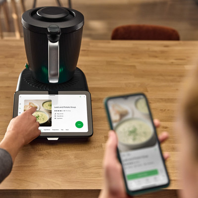 Le robot thermomix tm7 est sur un plan de travail et allumé. Sur l'écran on voit une recette de soupe sur la plateforme Cookidoo. En premier plan en flou, une personne a ouvert la même recette de soupe sur l'application mobile Cookidoo.