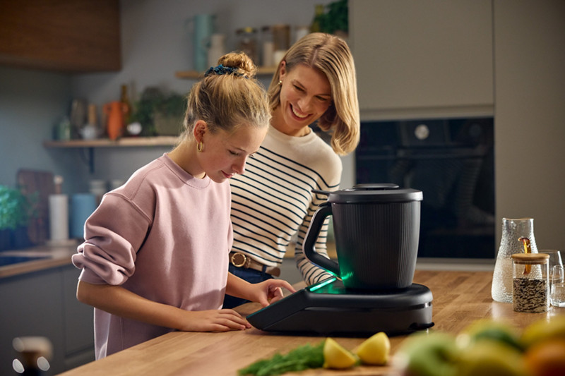 Une femme et sa fille cherche une recette sur le Thermomix, qui est placé au centre du plan de travail. 