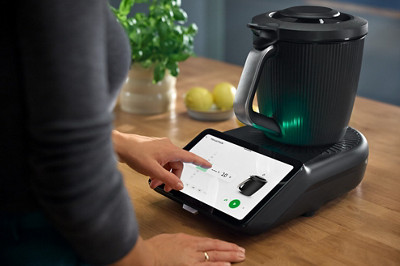 Le Robot multifonction Thermomix® TM7 est posé sur une table. L'écran du Thermomix est allumé et une dame lance une recette. 