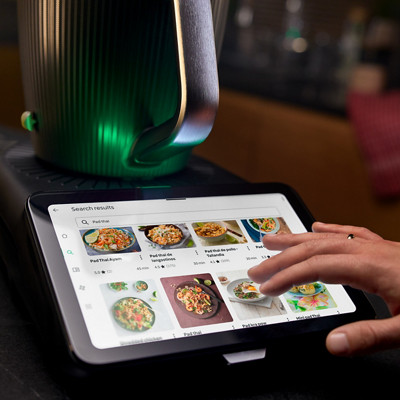 Le robot thermomix tm7 est sur un plan de travail et allumé. Sur l'écran on voit la plateforme Cookidoo proposant plusieurs recettes; 