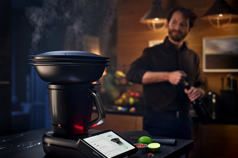 En premier plan, le Thermomix TM7 posé sur un plan de travail est en train de cuire des aliments à la vapeur avec le Varoma. En second plan, un homme débouche une bouteille.