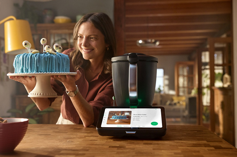 Le robot thermomix tm7 est sur un plan de travail, au centre et allumé. Sur l'écran, la plateforme Cookidoo montre une recette de gâteau. Une femme sur la gauche porte le gâteau de la photo, qu'elle a fait au robot Thermomix. 