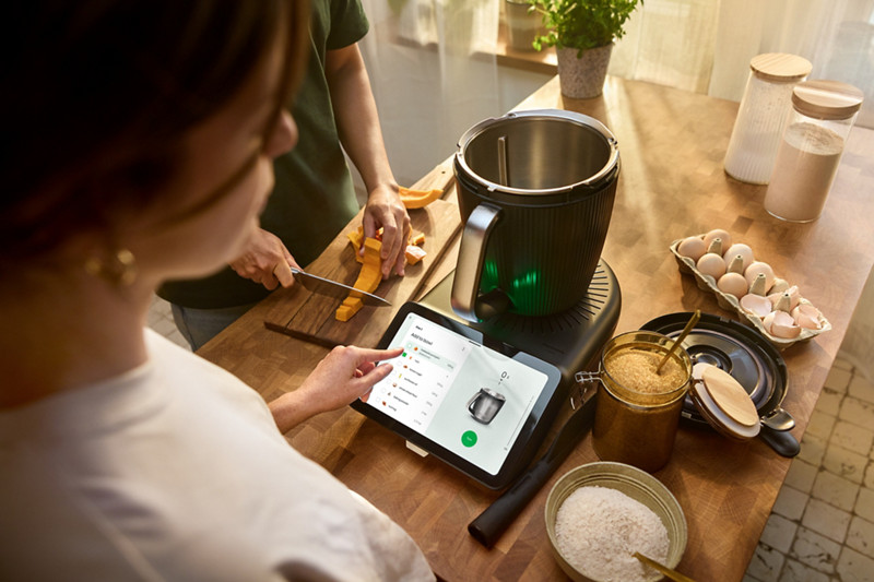 Le Thermomix TM7 placé au centre d'un plan de travail avec plusieurs aliments autour dont des oeufs, de la farine, du sucre... 