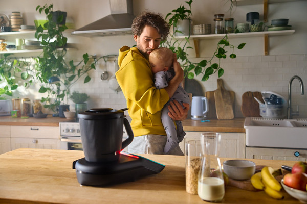 Un père porte son bébé et le Thermomix TM7 est posé devant lui sur un plan de travail