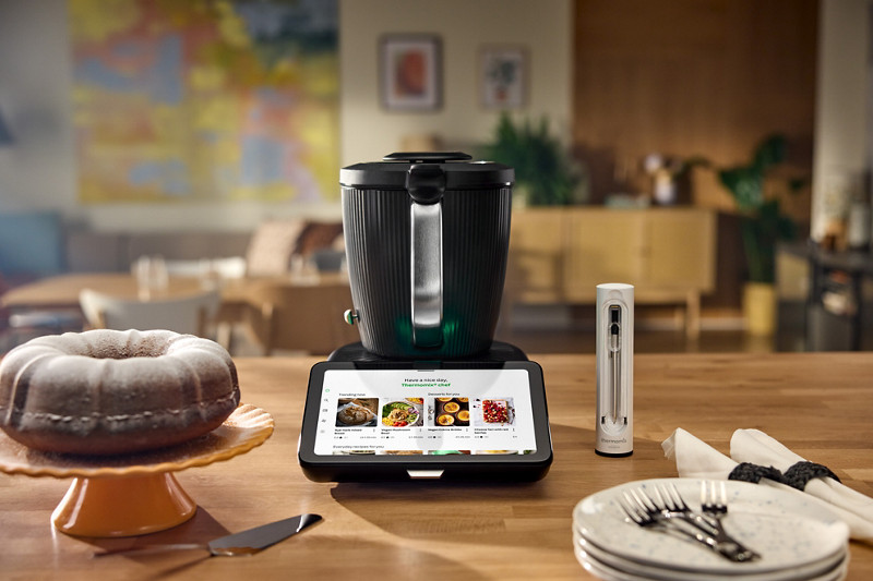 Le Thermomix TM7 placé au centre d'un plan de travail, avec sur la gauche un gateau marbré et à droite le Thermomix Sensor