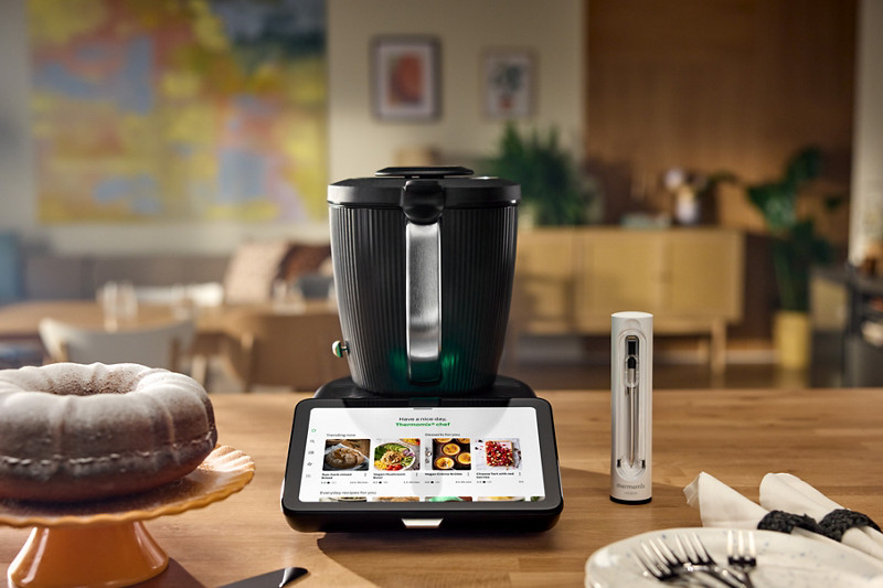 Le Thermomix TM7 placé au centre d'un plan de travail, avec sur la gauche un gateau marbré et à droite le Thermomix Sensor