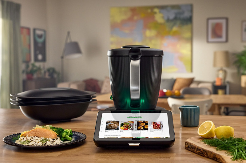 Le Thermomix est au centre d'un plan de travail avec l'écran allumé sur la plateforme de recette Cookidoo. A gauche, il y a le Varoma avec une assiette de saumon, riz, brocolis. A droite, il y a une planche à découper avec de l'aneth et du citron. 