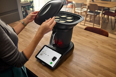 Le robot thermomix tm7 est sur un plan de travail, allumé. Une personne retire le couvercle du Varoma positionné sur le bol et on voit des yaourts à l'intérieur qui ont terminé de cuire.