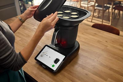 Le robot thermomix tm7 est sur un plan de travail, allumé. Une personne retire le couvercle du Varoma positionné sur le bol et on voit des yaourts à l'intérieur qui ont terminé de cuire.