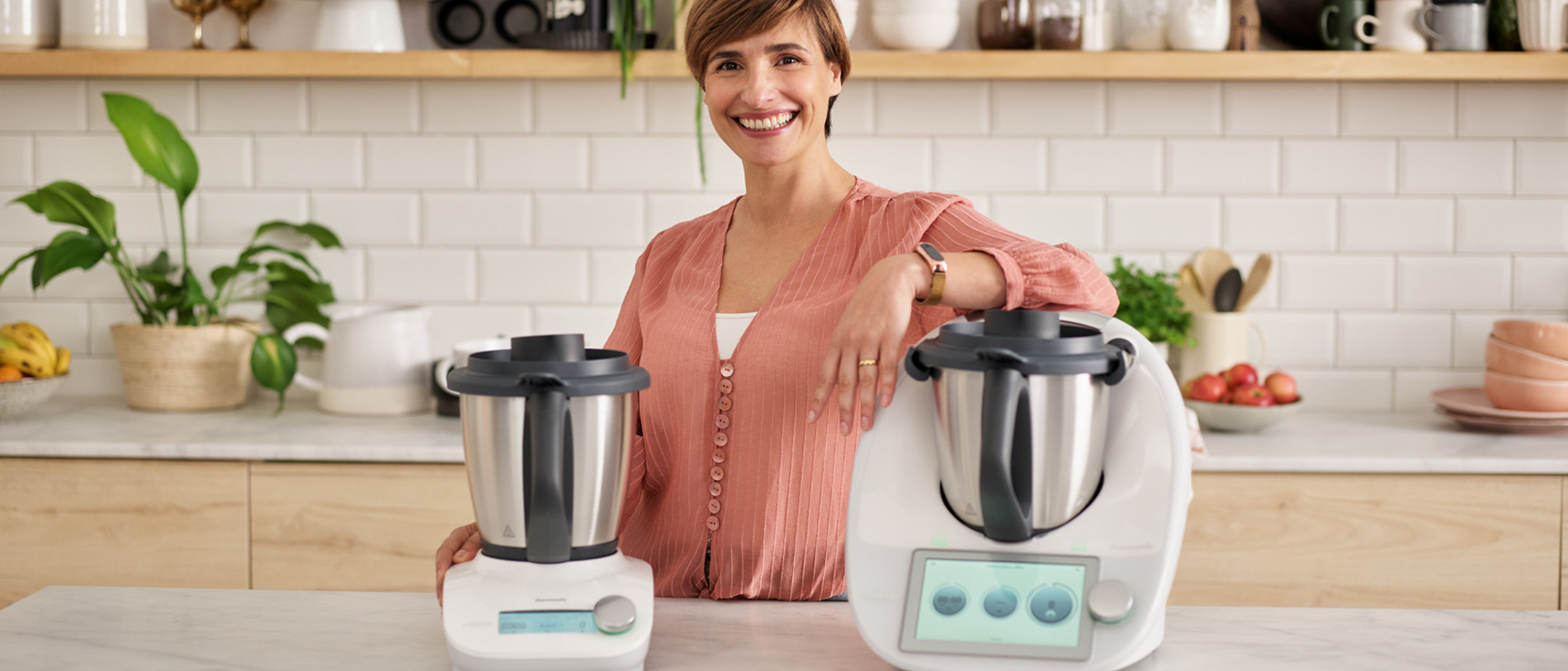Im Bild ist eine helle und moderne Küche abgebildet. Auf der Arbeitsplatte der Kücheninsel stehen der Thermomix TM6 und der Thermomix Friend in frontaler Ansicht. Eine Frau mit kurzen, bräunlichen Haaren und einer rosafarbenen Bluse lehnt mit einem Arm lächelnd auf dem Thermomix TM6.