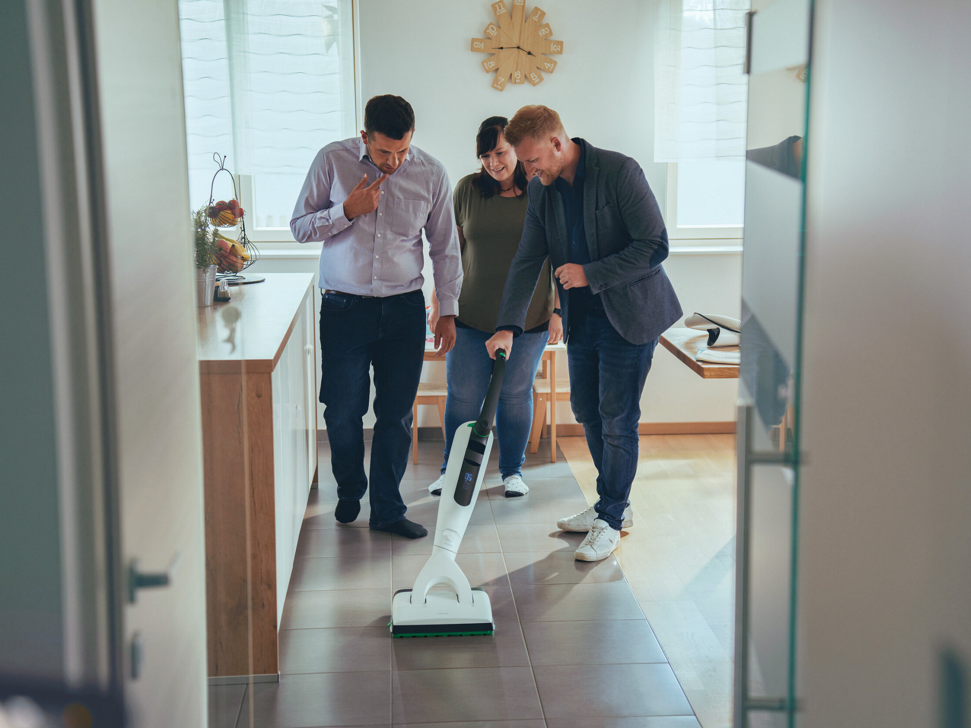 Démonstration du nettoyage du sol carrelé d’une cuisine avec l’aspirateur Kobold VK7, trois personnes présentes.