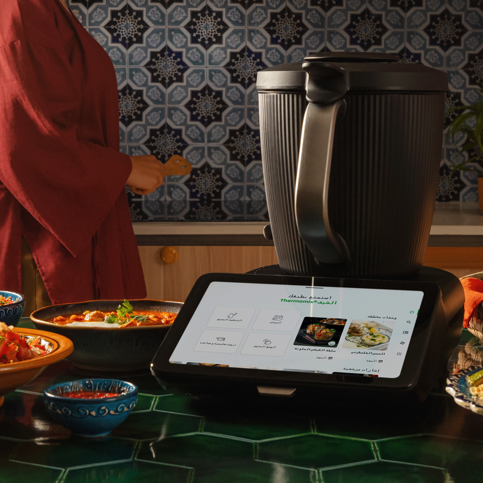 Thermomix® TM7 auf einer Küchenarbeitsplatte mit verschiedenen Gerichten, Brot und einer Person im Hintergrund.