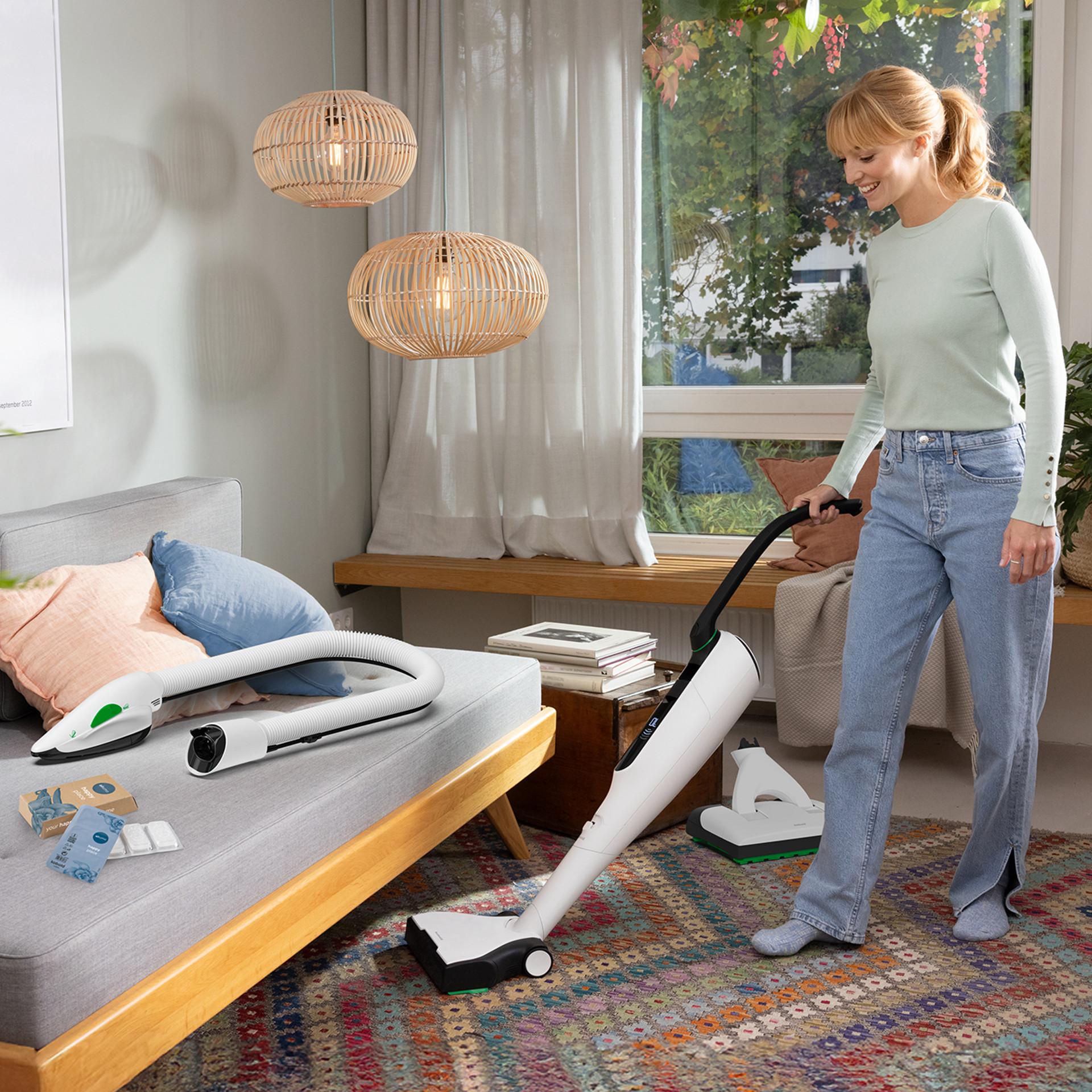 Frau reinigt das Wohnzimmer mit Kobold VK7 Akku-Staubsauger mit HD7 Hartbodendüse; Kobold PB7 Elektro-Polsterbürste liegt auf dem Sofa.