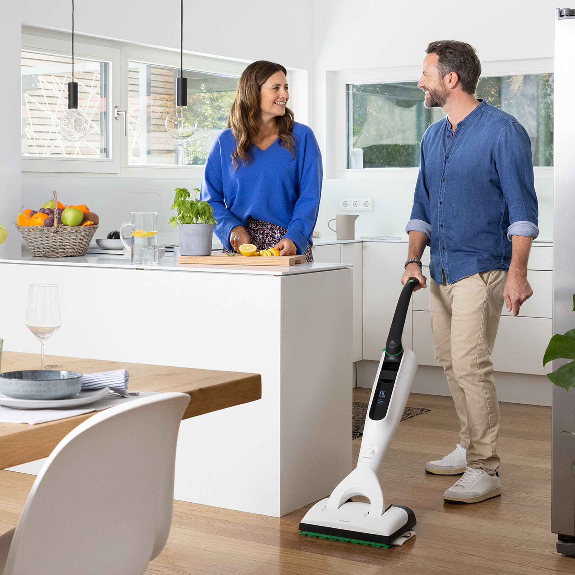 Ein Mann reinigt den Küchenboden mit einem Kobold VK7, während eine Frau am Tresen Essen zubereitet.