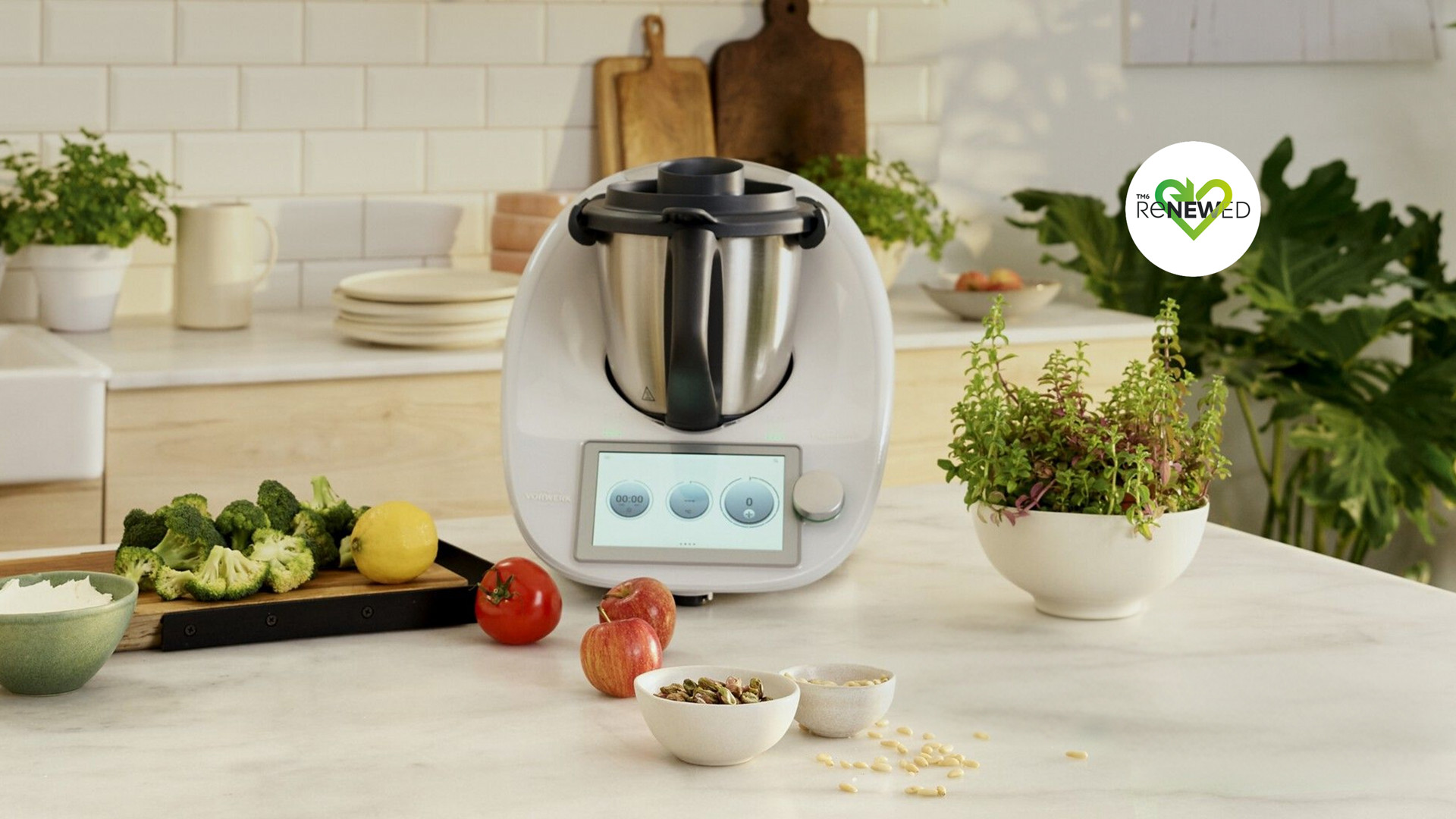 Thermomix® TM6 auf einer Küchenarbeitsplatte mit frischem Gemüse, Obst, Kräutern und Schalen zur Essenszubereitung.