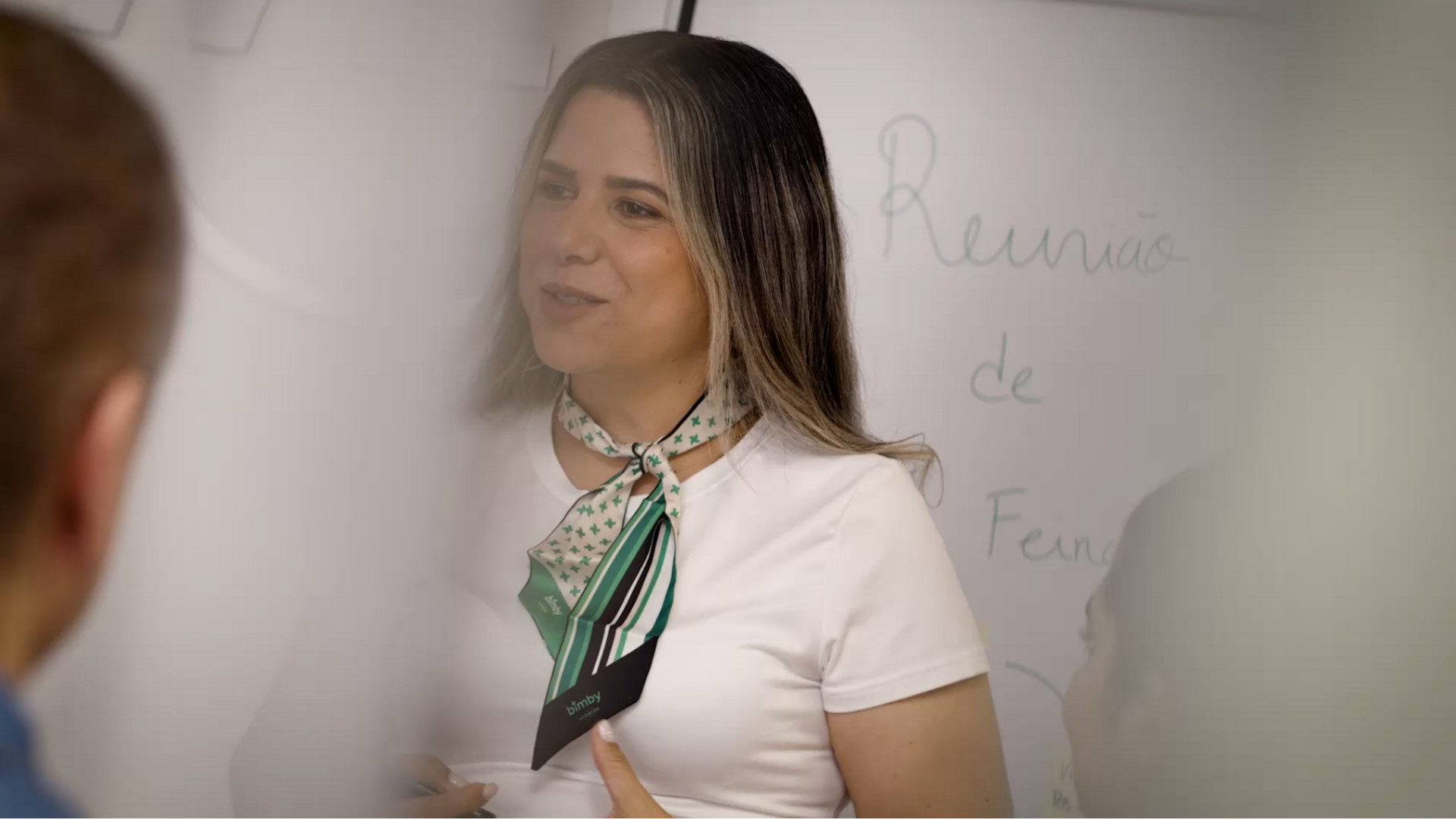 Mulher com lenço verde e branco Bimby falando diante de um quadro com escrita em português numa reunião.