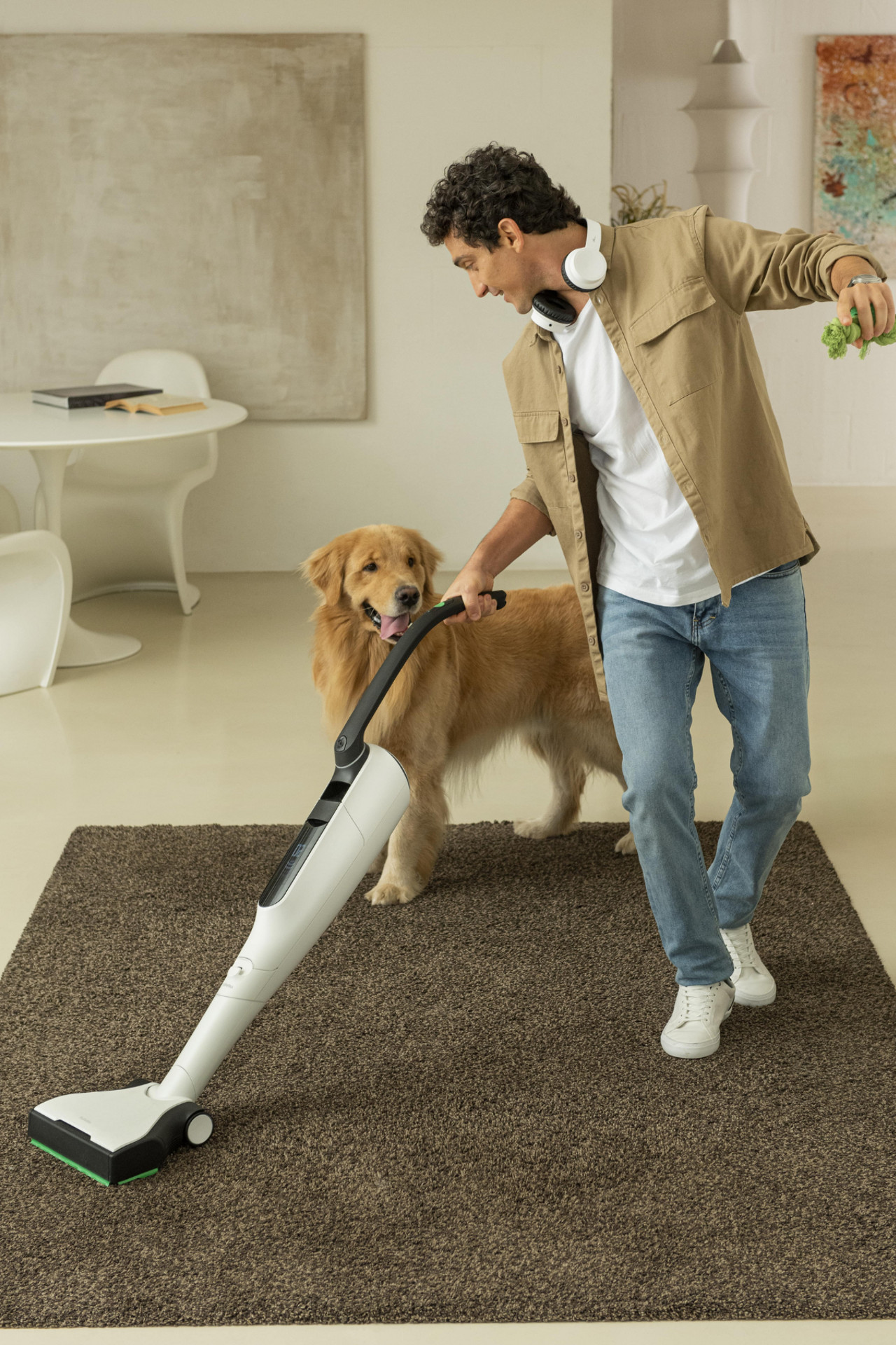 Uomo che aspira un tappeto accanto a un golden retriever con Folletto VK7s in un soggiorno moderno.