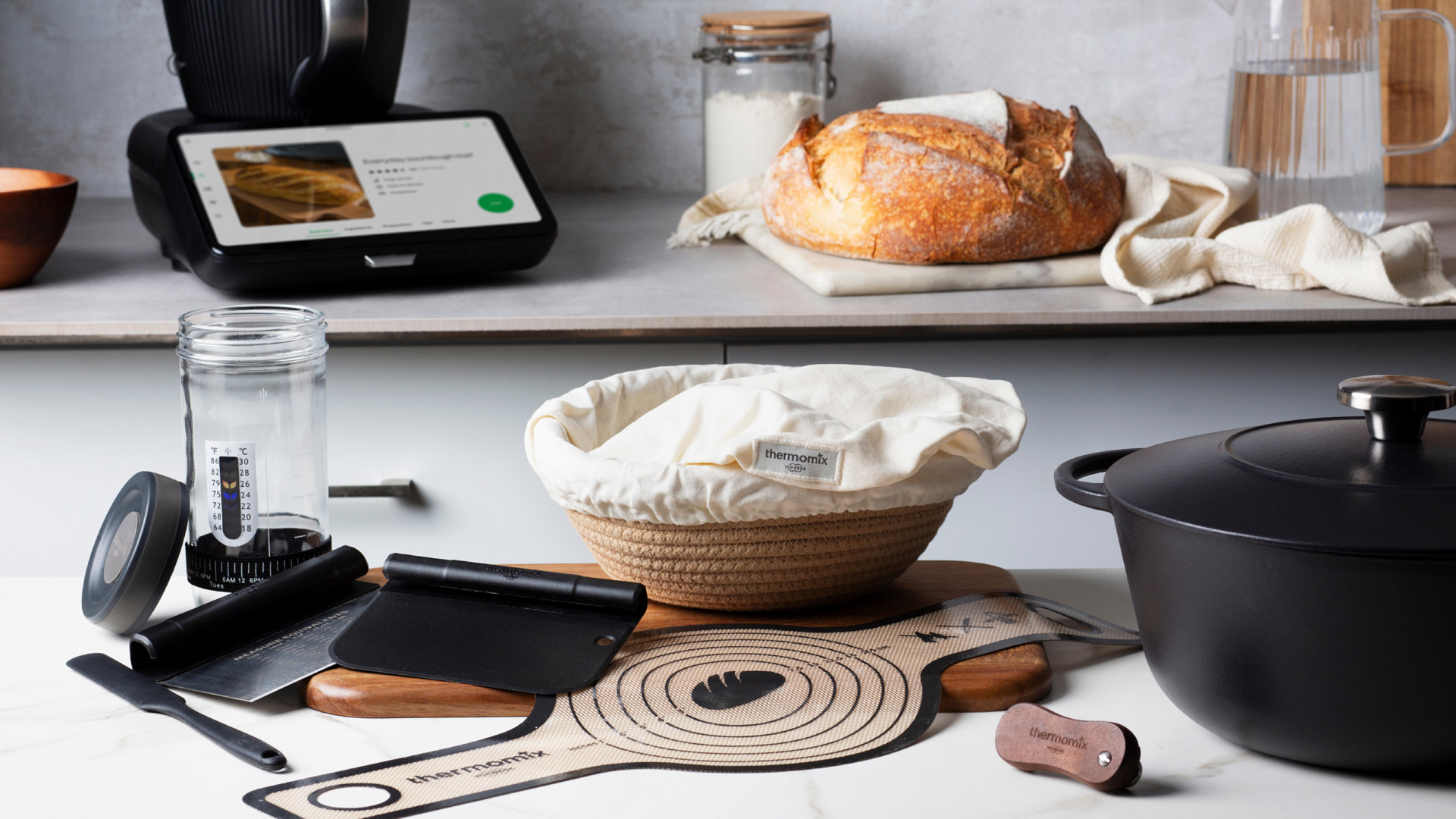 Küchenszene mit Thermomix® TM7, Backutensilien, Gärkorb, gusseisernem Topf und frisch gebackenem Brot.